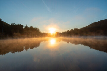 Morning sunrise over lake and forest, beautiful blue sky, sun and mist and reflection on the surface of the lake. Summer time.