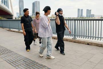 Obraz premium Group of diverse friends strolling along waterfront promenade with modern buildings in background, some wearing casual attire, engaging in lively conversation