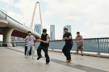 Group of young men dancing energetically beside modern bridge under clear sky capturing urban lifestyle and movement dynamics with background of city skyline and high-rise buildings