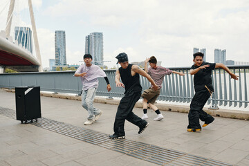 Four dancers performing dynamic hip-hop routine on urban bridge with cityscape in background creating energetic atmosphere with stylish moves and coordination