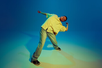 Lost in the music. Dynamic image for young man, hip-hop dancer making energetic performance against gradient blue background in neon light. Concept of street style, contemporary dance, entertainment