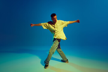 Obraz premium Cool guy, talented and artistic man energetically dancing hip-hop making dynamic performance against gradient blue background in neon light. Concept of street style, contemporary dance, entertainment
