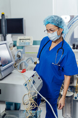 Medical professional monitoring equipment in a hospital setting. A healthcare worker in blue scrubs checks vital signs on a monitor