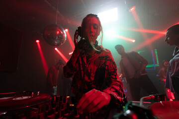 Short haired beautiful Dj woman standing behind the control panel, playing music and dancing with a...