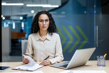 Hispanic businesswoman in office setting examining documents and using laptop. Professional and focused, showcasing concept of business and modern work environment.