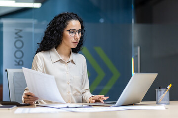 Hispanic businesswoman sits at office desk, analyzing documents and working on laptop. Professional attire conveys focus and efficiency in modern workplace environment.