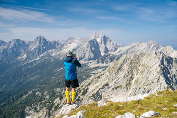Hiker captures the stunning mountain scenery on a smartphone, standing on a rocky outcrop with a backdrop of majestic peaks.  Perfect setting for outdoor photography