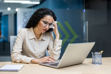 Hispanic businesswoman focused on laptop work, displaying concern in modern office setting. Professional attire and glasses emphasize dedication to task amidst working environment