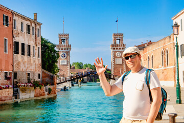 A cheerful traveler poses beside a picturesque canal with historic towers majestically in the...