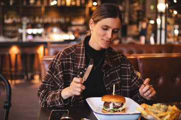 Woman eating a burger in a restaurant, knife and fork prepared. Cheat meal, fast food. Portrait of a young lovely hungry woman sitting in the fast junk food restaurant cafe. Unhealthy eating. 
