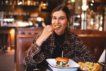 Fast food lover. Happy lady eating burger and french fries, biting potatoes and looking camera, sitting in restaurant. Playful woman holding french fries, burger in plate and tasty. Cheat meals.
