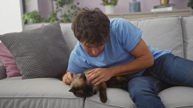 A young hispanic adult man lovingly pets a siamese cat on a gray couch in a cozy living room, evoking a sense of home warmth.