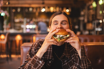 Close-up portrait of a woman in a fast food restaurant eating a hamburger. Hungry woman eating a burger sitting in cafe. Girl bite bbq burger, look joyful. Cheat meal day concept.