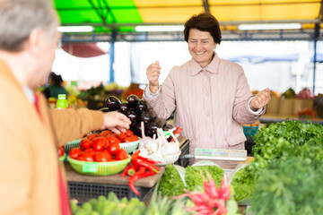 Smiling elderly female seller telling prices for fresh vegetables at local bazaar