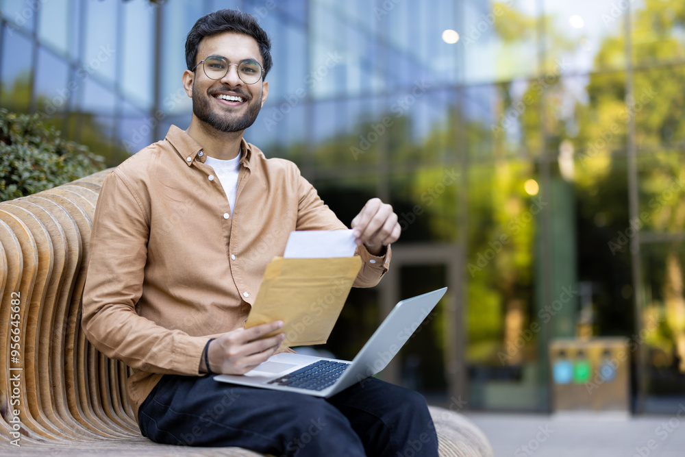 Wall mural young adult wearing glasses holds envelope with smile, seated on outdoor bench with laptop open. bri