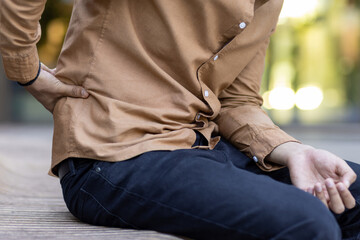 Individual seated on bench outdoors showing signs of discomfort due to lower back pain. Casual...