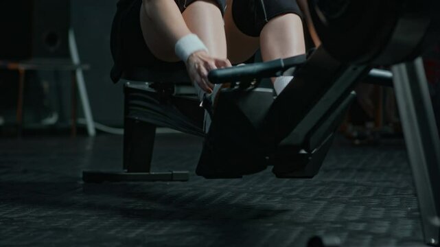 Tilt up shot of young sportswoman pulling laces of her sneakers and starting to exercise on rowing machine