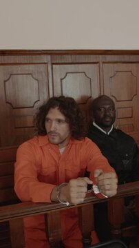 Vertical shot of Caucasian young male criminal wearing orange jumpsuit and handcuffs sitting near African American policeman at court session