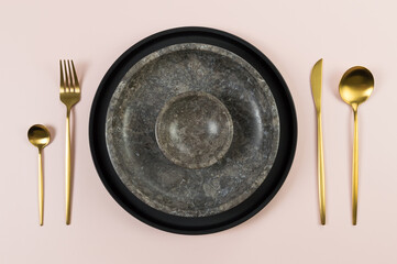 Top view of brown marble plate and bowl, black plate and gold cutlery on pink background. Food...