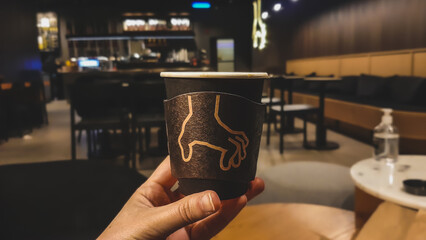 Woman holding a takeaway coffee cup with a grabbing hand sign on it, in a coffee shop