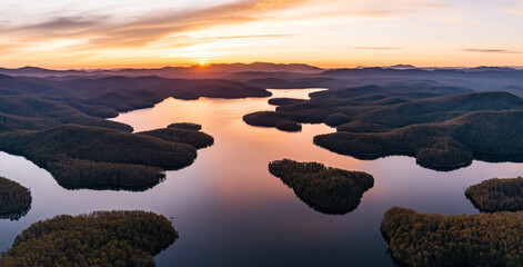 Aerial view of a serene lake with numerous forested islands and peninsulas during a sunset, creating a tranquil and picturesque landscape.