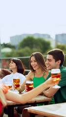 Young people sitting on terrace of urban pub, and clinking their beer mugs, celebrating meeting after long years. Selective focus. Concept of Friday mood, celebration, lunch break and Oktoberfest.
