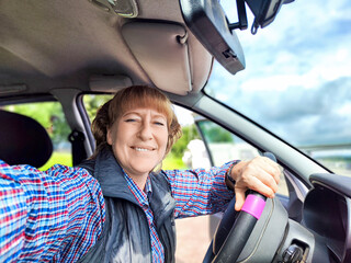 Middle aged woman in a car taking selfie while enjoying a drive. Female mature driver posing inside...