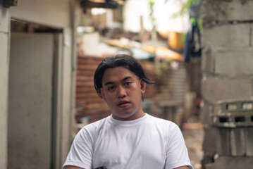 A young asian man looking at the camera with a gloomy face full of apathy. Feeling down with financial problems. Slum area background.