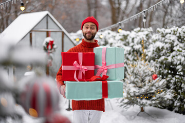 Cheerful man in red sweater and hat carries gift boxes at snowy backyard. Concept of happy winter time, Thanks giving day and Valentine holiday