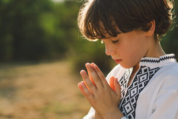 The child stands with hands together in a prayer pose, wearing a traditional embroidered shirt, amidst a serene outdoor setting with soft sunlight filtering through trees.