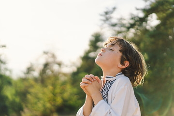 The child stands with hands together in a prayer pose, wearing a traditional embroidered shirt,...