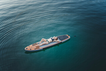 Young cheerful woman relaxing lying down on a sup surf board on the water in a mountain lake