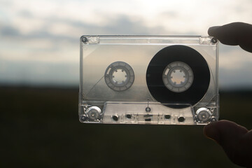 man holding transparent audio cassette between two male fingers on blue sky and field background. magnetic tape. old technology.