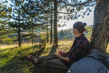 Fototapeta premium Young female hiker sitting down to rest on the grass during a summer hiking in pine woodland