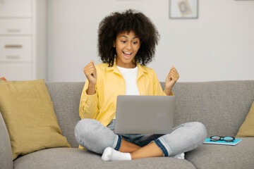 Excited black lady sitting on couch with laptop, shaking fists, gesturing YES and celebrating success while sitting on sofa at home