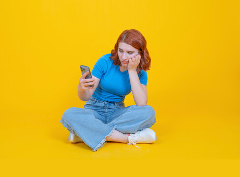 Bored young caucasian woman, full body view sitting legs crossed bored young caucasian woman holding looking at smartphone screen over yellow studio background. Waiting important call or message.