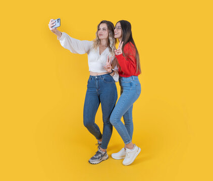 Doing selfie shot, full body length excited beautiful attractive two young women doing selfie shot. Best friends wearing casual denim jean taking photo on phone show victory sign over yellow color.