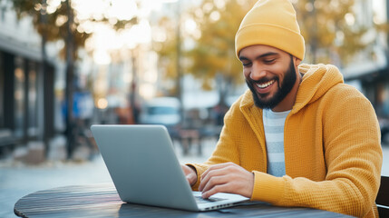 A man in a yellow hoodie and beanie works on his laptop in a vibrant city square, capturing the...