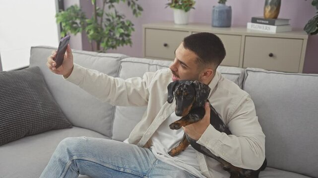Young hispanic man having a video call at home while holding a teckel dog in a modern living room.