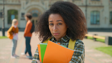 Sad upset stressed disappointed African American student girl female woman lady teen teenager standing alone in city outdoors university college high school suffer bullying talking students background