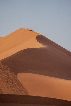 Wind blowing sand over the top of a giant sand dune at sunrise in Sossusvlei, Namibia