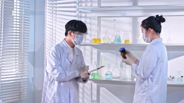 Scene in a laboratory with female researcher holding unlabeled vial of solution and discussing with male researcher taking notes and some experimental equipments on the shell.