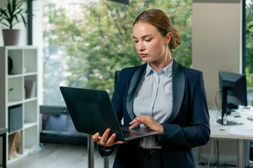 Fototapeta premium in a modern office with a large window beautiful female worker stands in a blue suit with a laptop in her hand looking through the work plan for the month