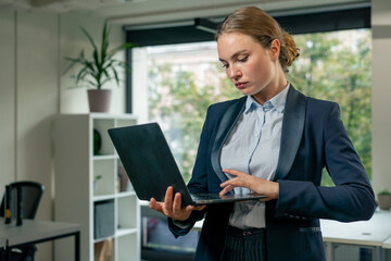 in a modern office with a large window beautiful female worker stands in a blue suit with a laptop in her hand looking through the work plan for the month