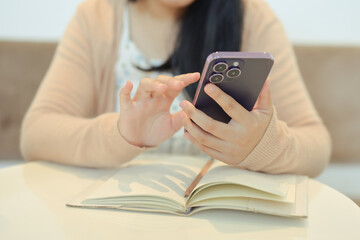 Young woman in casual dressed using smartphone on wooden table next to a notebook