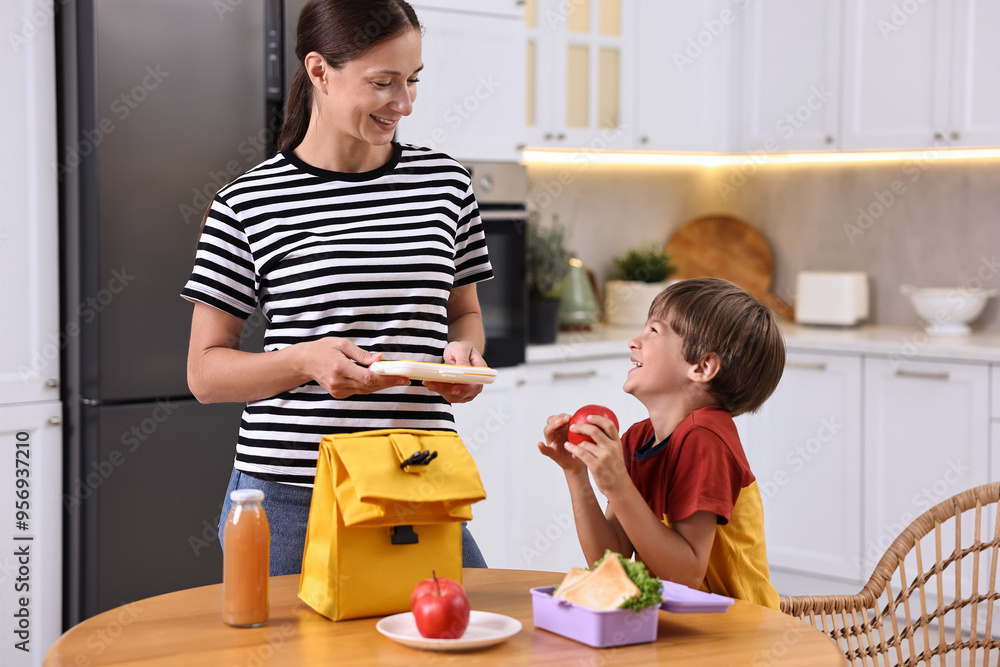 Poster Smiling mother and her cute son preparing school lunch box with healthy food at wooden table in kitchen