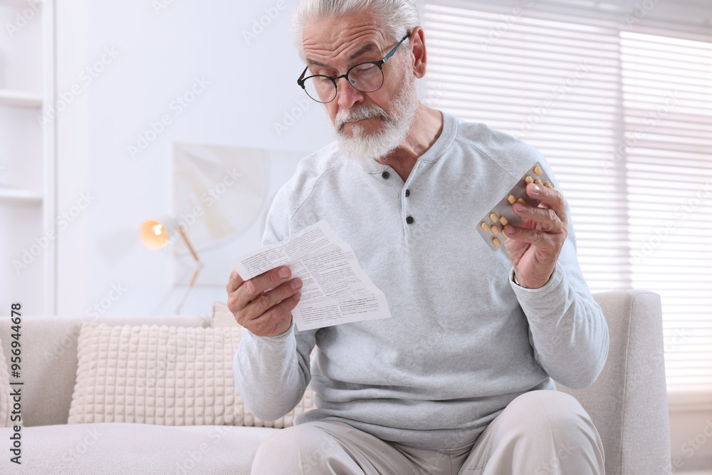 Wall mural Senior man with pills reading medicine instruction on sofa at home