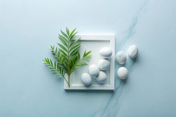 A white frame with white round stones inside, a green branch is on the left side.  There are four round white stones on the right side of the frame.  The background is a light blue marble surface.