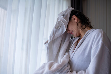 Beautiful woman with wet hair is drying their face with a towel wearing a white bathrobe and are...