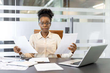 Fototapeta premium African American businesswoman reviewing important documents at work desk. Professional setting with laptop, creating focused and determined atmosphere. Represents modern business, office work